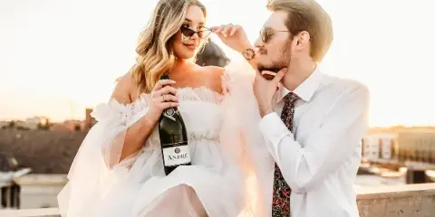 A bride in a white off-the-shoulder gown and a groom in a white shirt and floral tie pose together on a rooftop at sunset, with the bride holding a champagne bottle.