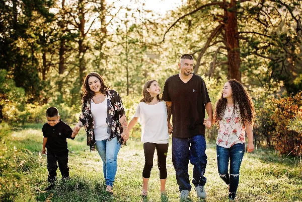 A family of five walking hand-in-hand through a sunlit wooded area during a professional outdoor portrait session.