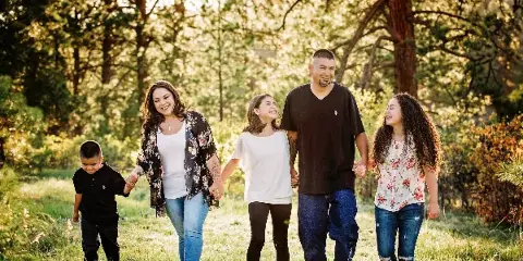 A family of five walking hand-in-hand through a sunlit wooded area during a professional outdoor portrait session.