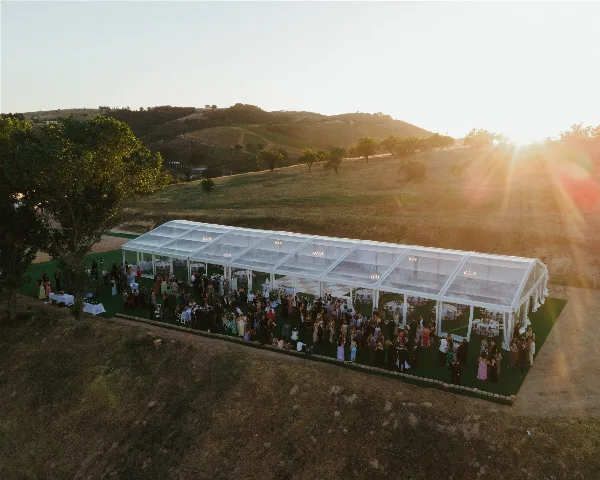 Aerial view of a large clear-frame wedding tent set up on a grassy hillside with guests and tables inside.