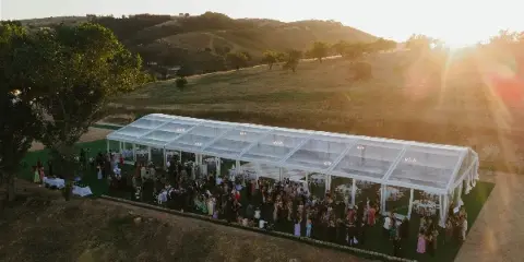 Aerial view of a large clear-frame wedding tent set up on a grassy hillside with guests and tables inside.