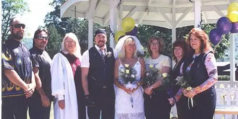 A wedding party, including the bride, groom, and officiant, posing together outdoors after a ceremony.
