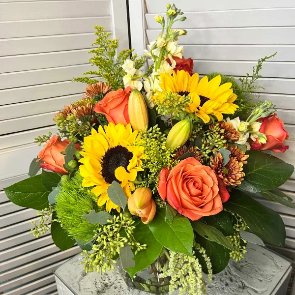 A mixed floral bouquet containing sunflowers, orange roses, and lilies arranged in a clear glass vase.