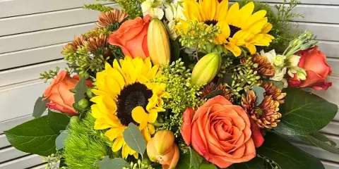 A mixed floral bouquet containing sunflowers, orange roses, and lilies arranged in a clear glass vase.