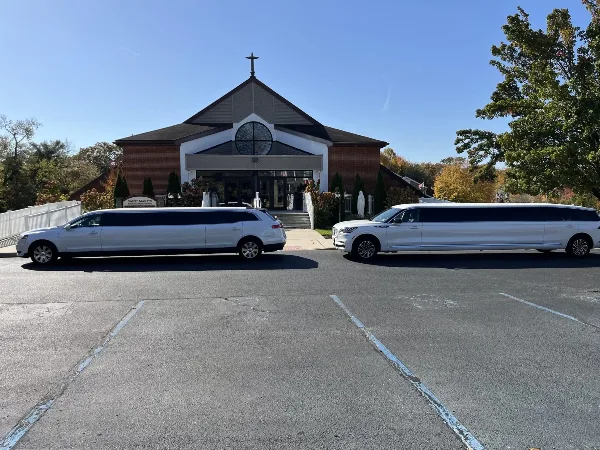 Two white stretch limousines parked in a lot in front of a brick church building.