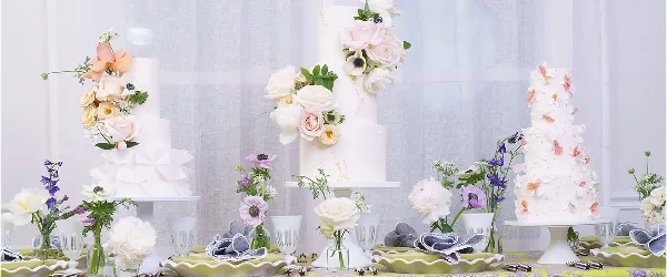 Three multi-tiered white wedding cakes decorated with fresh flowers displayed on a table with floral arrangements.