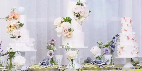 Three multi-tiered white wedding cakes decorated with fresh flowers displayed on a table with floral arrangements.