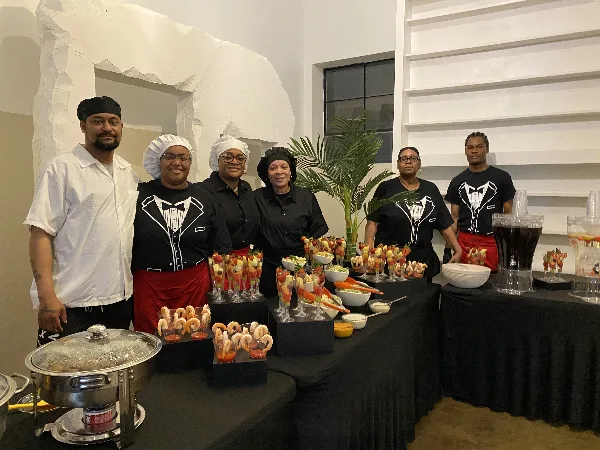 Catering staff in uniform standing behind a buffet table with prepared food and beverage dispensers.