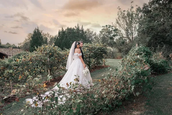 A bride and groom standing together in a garden with lush greenery and a stone bridge in the background.