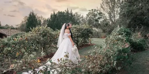 A bride and groom standing together in a garden with lush greenery and a stone bridge in the background.
