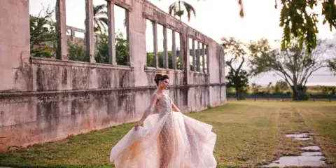 A bride in a flowing white wedding dress posing outdoors near a historic stone structure.