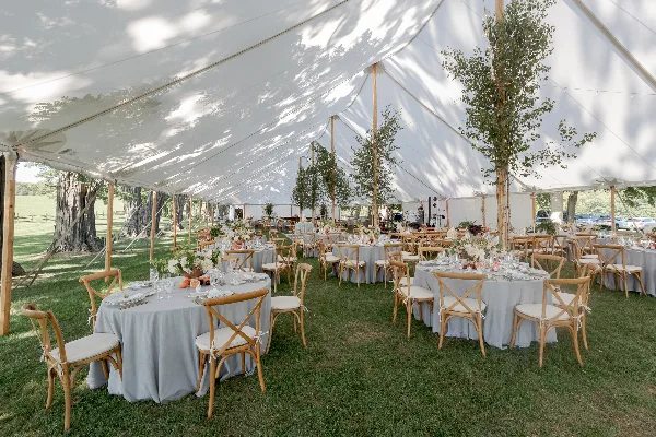 Interior view of a large white event tent set up on a grassy field, featuring multiple round tables with grey linens, wooden chairs, and floral centerpieces.
