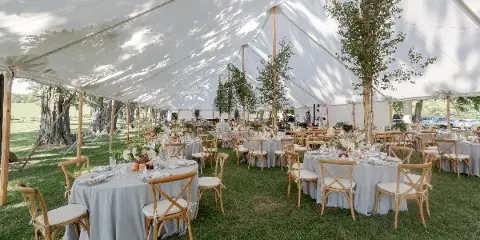 Interior view of a large white event tent set up on a grassy field, featuring multiple round tables with grey linens, wooden chairs, and floral centerpieces.