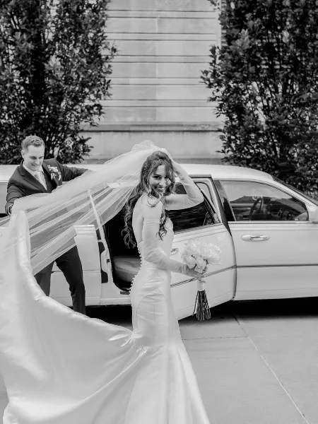 A bride in a white gown and veil steps out of a white limousine while holding a bouquet, with the groom smiling in the background.