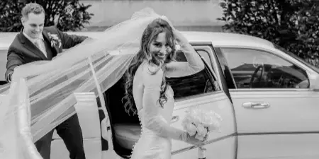 A bride in a white gown and veil steps out of a white limousine while holding a bouquet, with the groom smiling in the background.