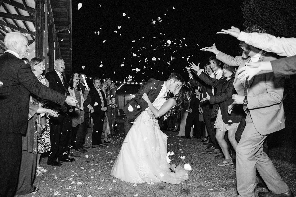 A black and white photo of a bride and groom walking through a line of guests who are throwing flower petals at them.