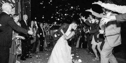 A black and white photo of a bride and groom walking through a line of guests who are throwing flower petals at them.