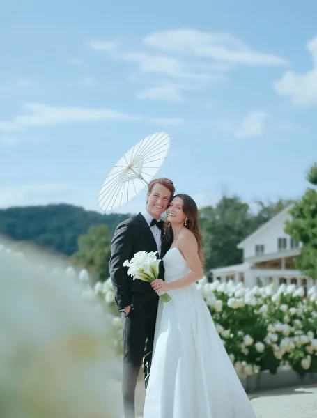 A bride and groom posing outdoors with a white parasol and bouquet, set against a backdrop of a white building and blue sky.