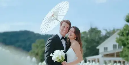A bride and groom posing outdoors with a white parasol and bouquet, set against a backdrop of a white building and blue sky.