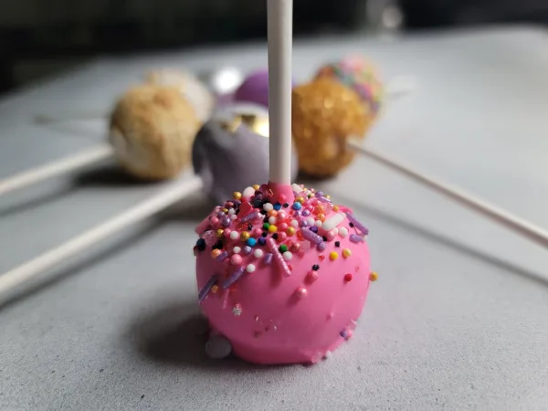 A close-up of a pink cake pop covered in colorful sprinkles, with other decorated cake pops in the background.