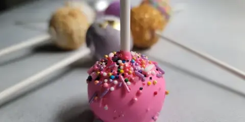 A close-up of a pink cake pop covered in colorful sprinkles, with other decorated cake pops in the background.