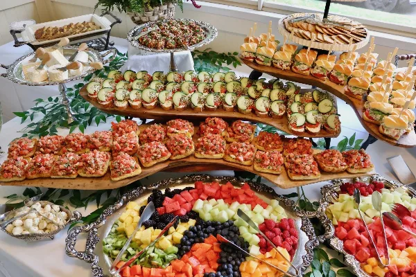 A catering table set with a tiered display of bruschetta, cucumber appetizers, and a large silver platter of assorted fresh fruit.