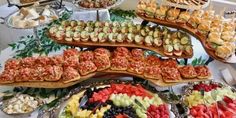 A catering table set with a tiered display of bruschetta, cucumber appetizers, and a large silver platter of assorted fresh fruit.