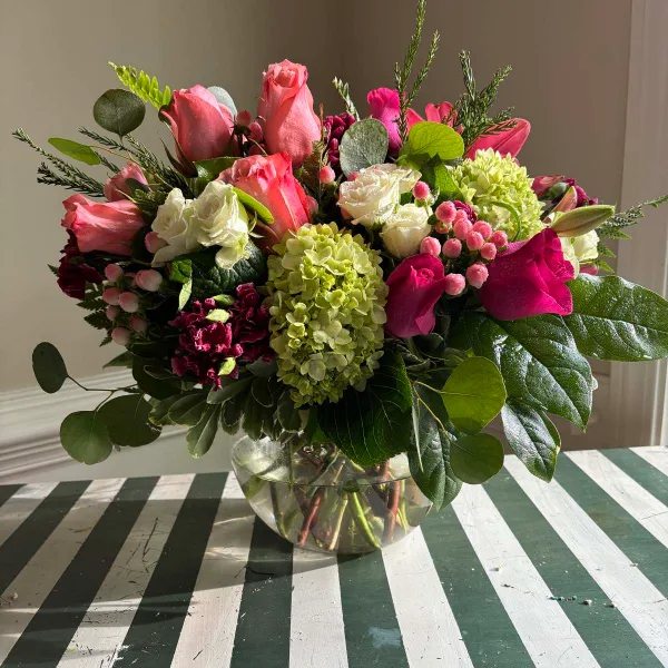 A mixed floral arrangement with pink and white roses, green hydrangeas, and eucalyptus in a clear glass vase on a striped table.