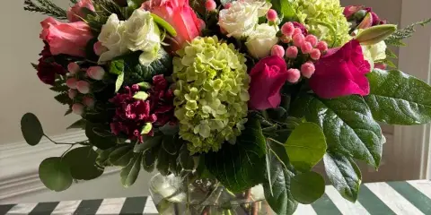 A mixed floral arrangement with pink and white roses, green hydrangeas, and eucalyptus in a clear glass vase on a striped table.
