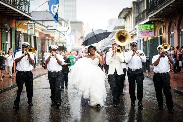 A bride and groom walking in a second line parade in New Orleans, surrounded by a brass band and guests.