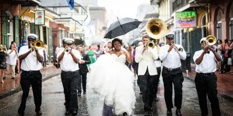 A bride and groom walking in a second line parade in New Orleans, surrounded by a brass band and guests.