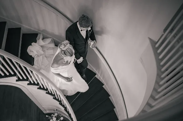 A black and white photograph of a bride and groom standing on a curved staircase, holding hands and looking down.