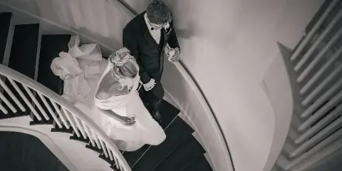 A black and white photograph of a bride and groom standing on a curved staircase, holding hands and looking down.