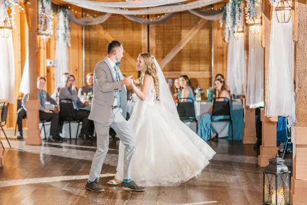 A bride and groom dancing at a wedding reception inside a rustic wooden barn venue.