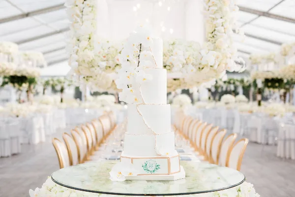 A tall, white, four-tiered wedding cake decorated with white orchids and gold accents, displayed on a glass table in a wedding venue.