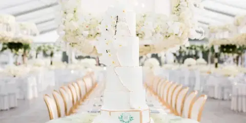 A tall, white, four-tiered wedding cake decorated with white orchids and gold accents, displayed on a glass table in a wedding venue.