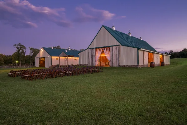 Exterior view of a white barn wedding venue with green roof and open doors at dusk, featuring rows of wooden chairs set up on a grassy lawn.