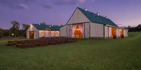 Exterior view of a white barn wedding venue with green roof and open doors at dusk, featuring rows of wooden chairs set up on a grassy lawn.