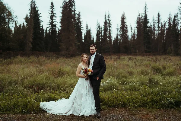 A bride and groom standing together in a grassy field with a backdrop of tall evergreen trees.