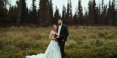 A bride and groom standing together in a grassy field with a backdrop of tall evergreen trees.
