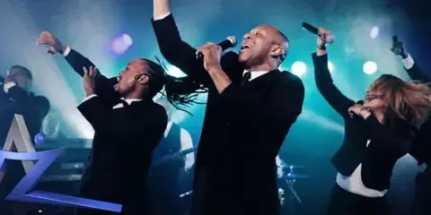 A live band performing on stage with multiple vocalists holding microphones under blue stage lighting.