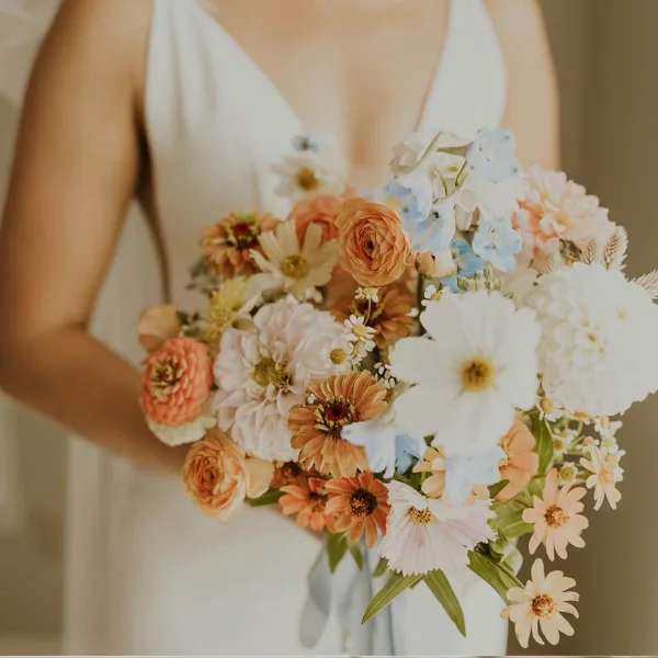 A bride in a white dress holding a large, colorful bouquet of flowers including ranunculus, daisies, and delphinium.