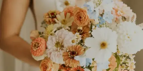 A bride in a white dress holding a large, colorful bouquet of flowers including ranunculus, daisies, and delphinium.