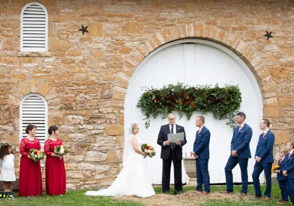 A wedding ceremony in progress with a bride, groom, and officiant standing before a rustic limestone barn with a white arched door.
