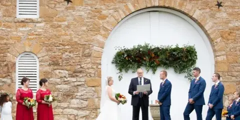A wedding ceremony in progress with a bride, groom, and officiant standing before a rustic limestone barn with a white arched door.