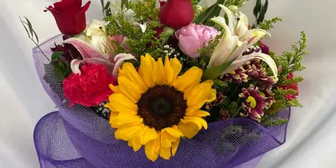 A mixed floral bouquet containing a sunflower, red roses, and lilies, wrapped in purple mesh.