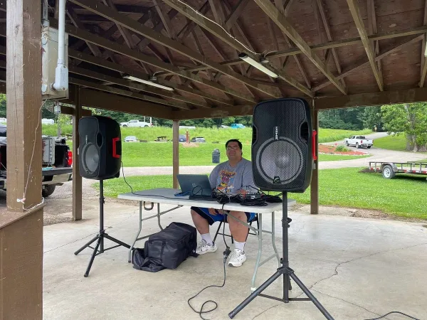 A DJ performing at an outdoor pavilion with a laptop and large speakers.