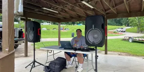 A DJ performing at an outdoor pavilion with a laptop and large speakers.