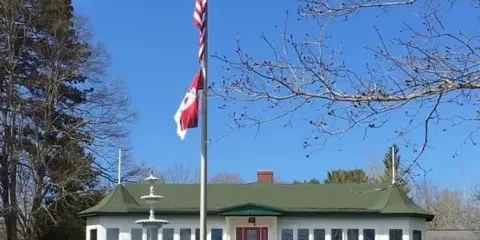 Exterior view of the Waldo County Shrine Club building with a green roof, white siding, red front door, and a flagpole with the American flag and a red flag.