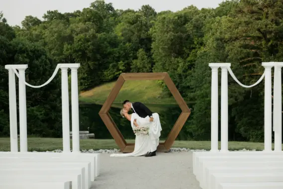 A bride and groom sharing a kiss under a wooden hexagonal arbor at an outdoor wedding venue.